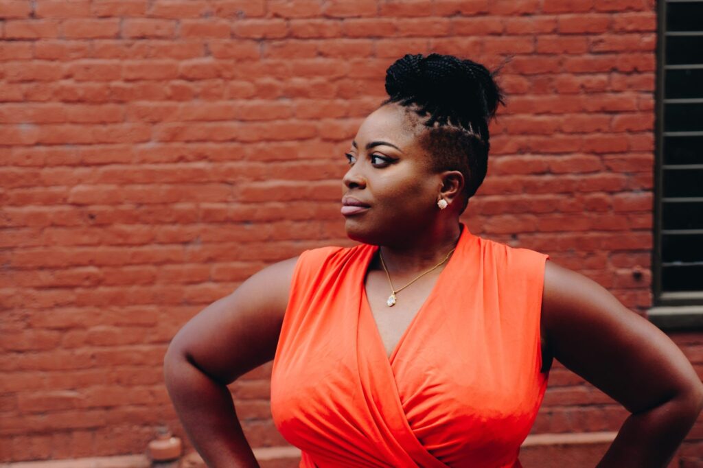 Dark complexioned woman of color with a short haircut, in an orange dress and looking into the distance in front of a brick wall.